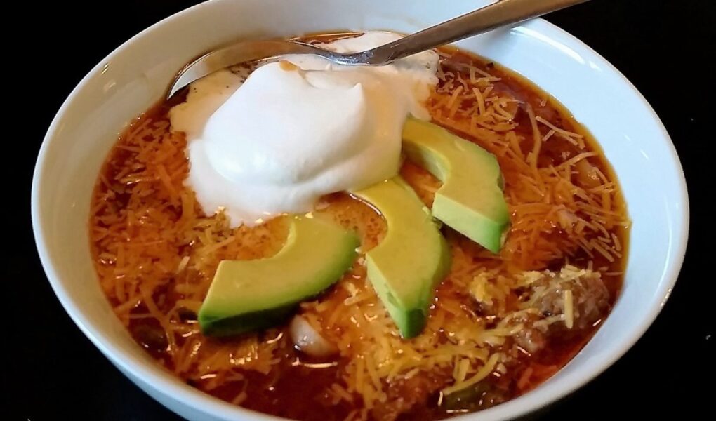 Chili served in white bowl, topped with 3 avocado slices and scoop of sour cream