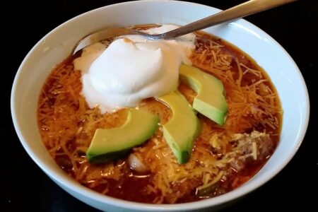 Chili served in white bowl, topped with 3 avocado slices and scoop of sour cream